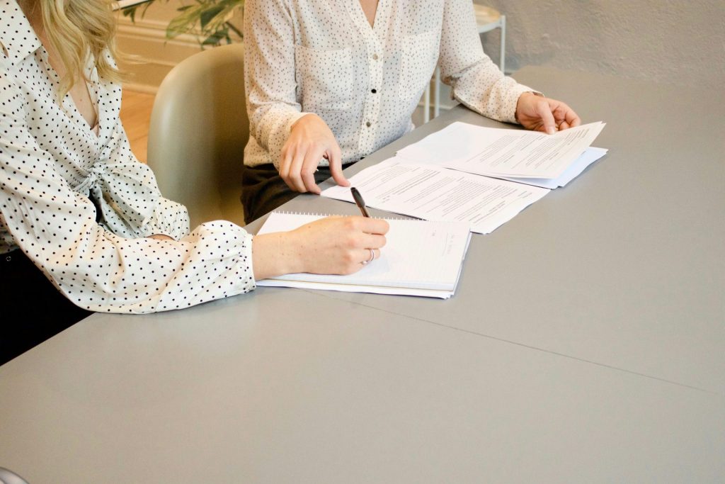 Eine Frau unterschreibt auf weißem Druckerpapier neben einer Frau, die im Begriff ist, die Dokumente zu berühren.